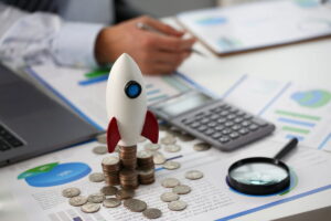 Modelo de foguete sobre pilhas de moedas, gráficos financeiros ao fundo, simbolizando sistema de controle financeiro.