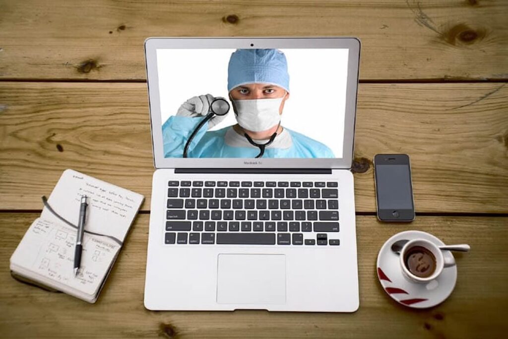 Médico em tela de laptop, simbolizando plataforma de telemedicina, com estetoscópio, caderno e celular ao lado.
