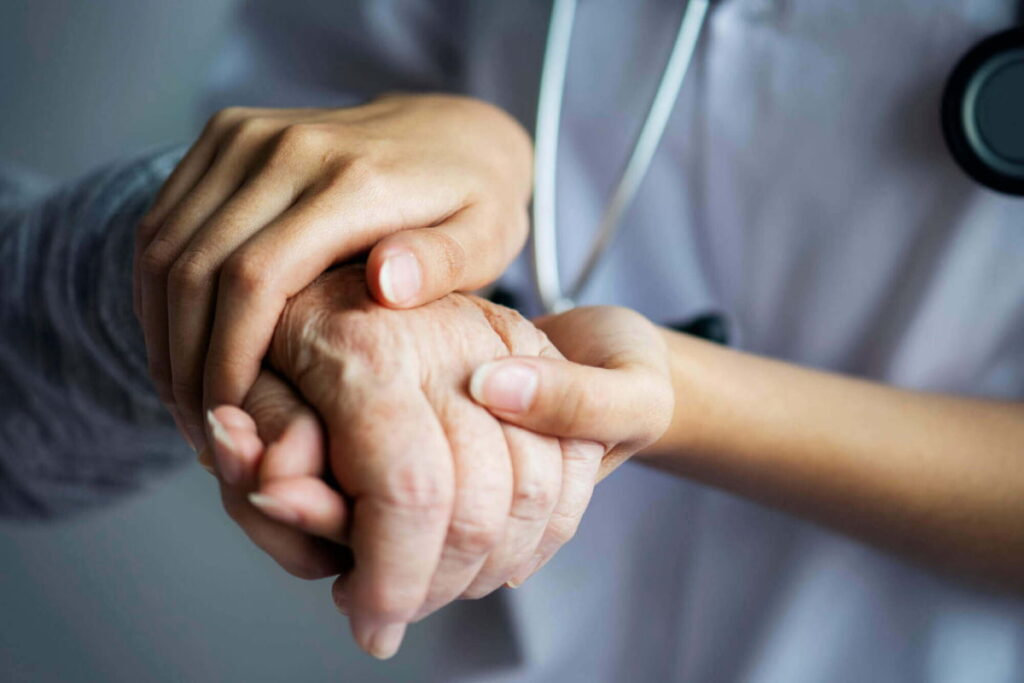 Mãos de profissional de saúde segurando a mão de um paciente idoso, simbolizando o atendimento preferencial na telemedicina.