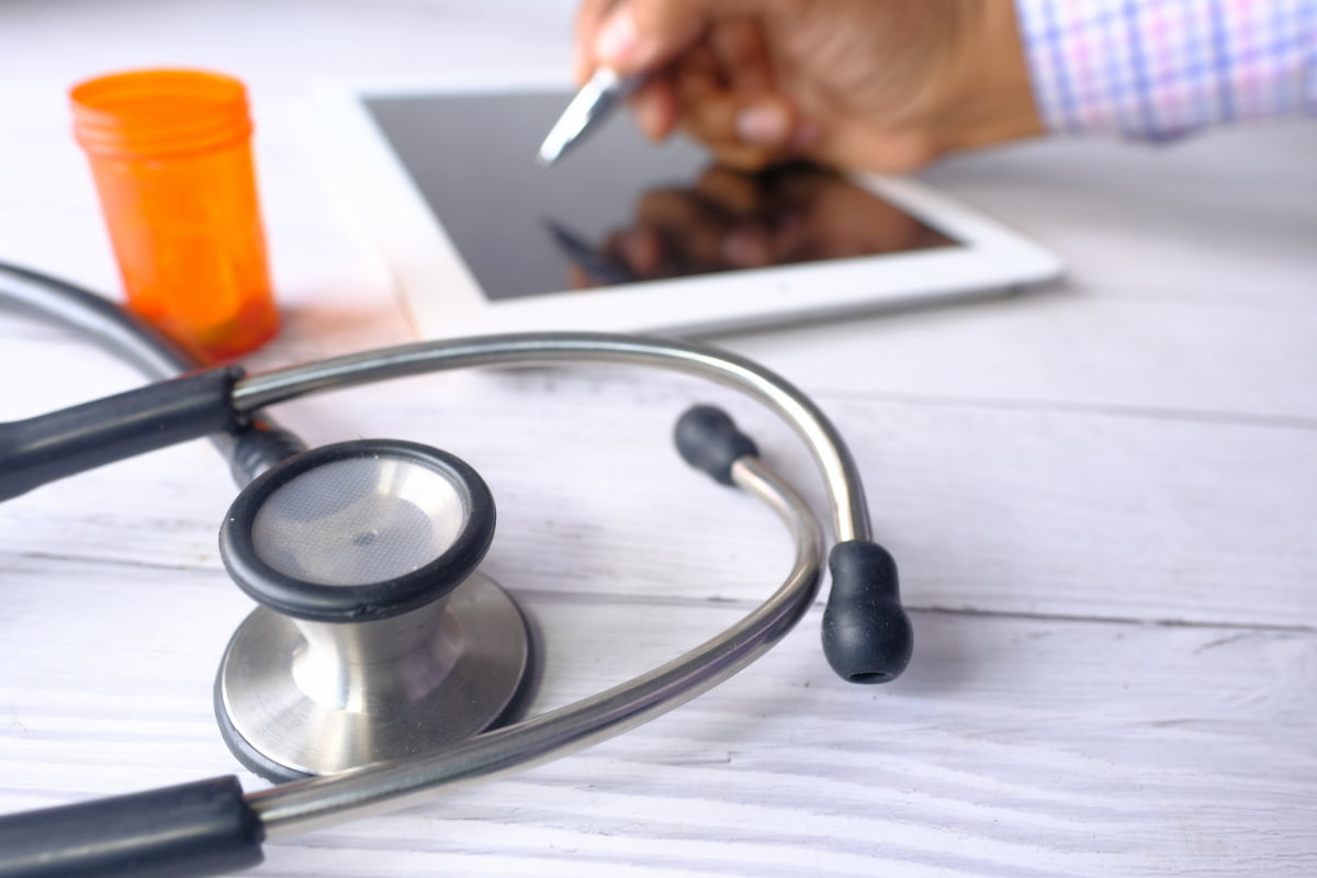 Mão com caneta sobre tablet, estetoscópio e remédios ao lado, simbolizando a MEMED Prescrição Digital.