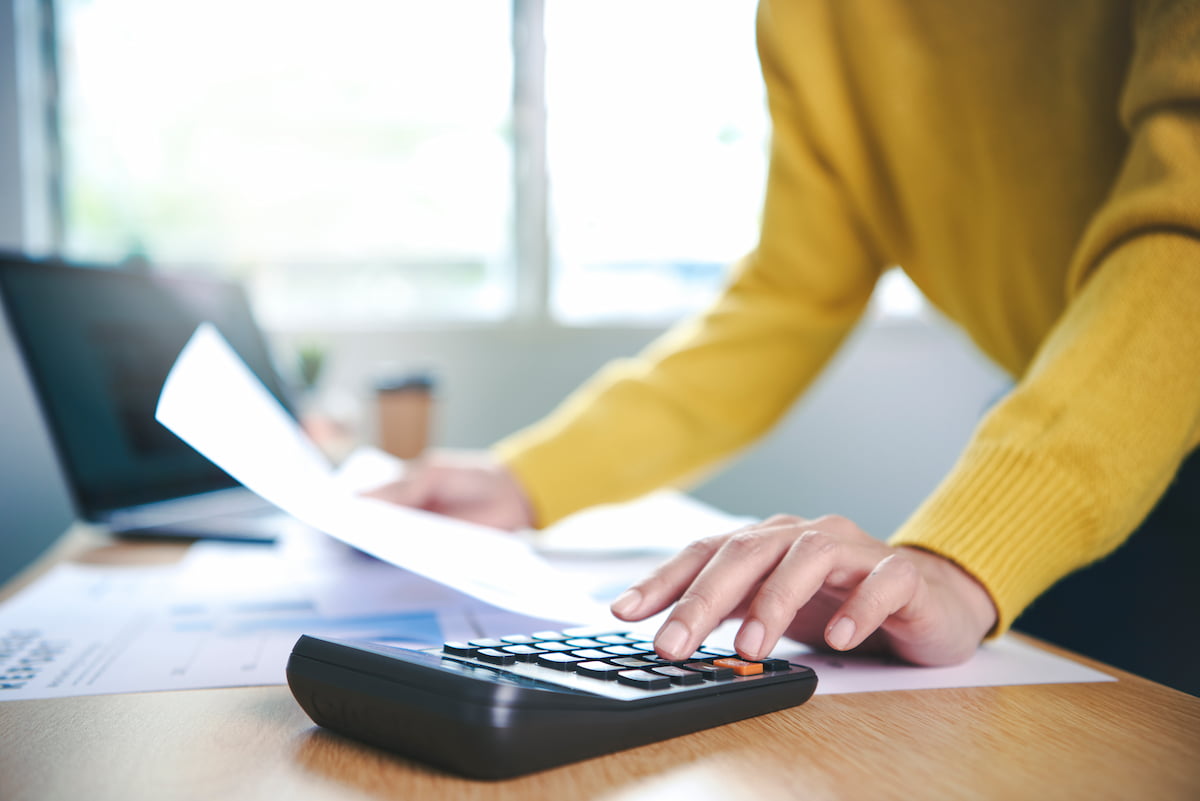 Pessoa utilizando calculadora para gerenciar cobrança recorrente, com papéis e laptop ao fundo.