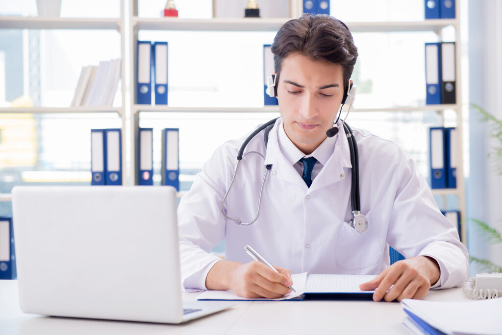 Médico de jaleco e com estetoscópio colocado sobre o ombro, realizando anotações em frente a um computador.