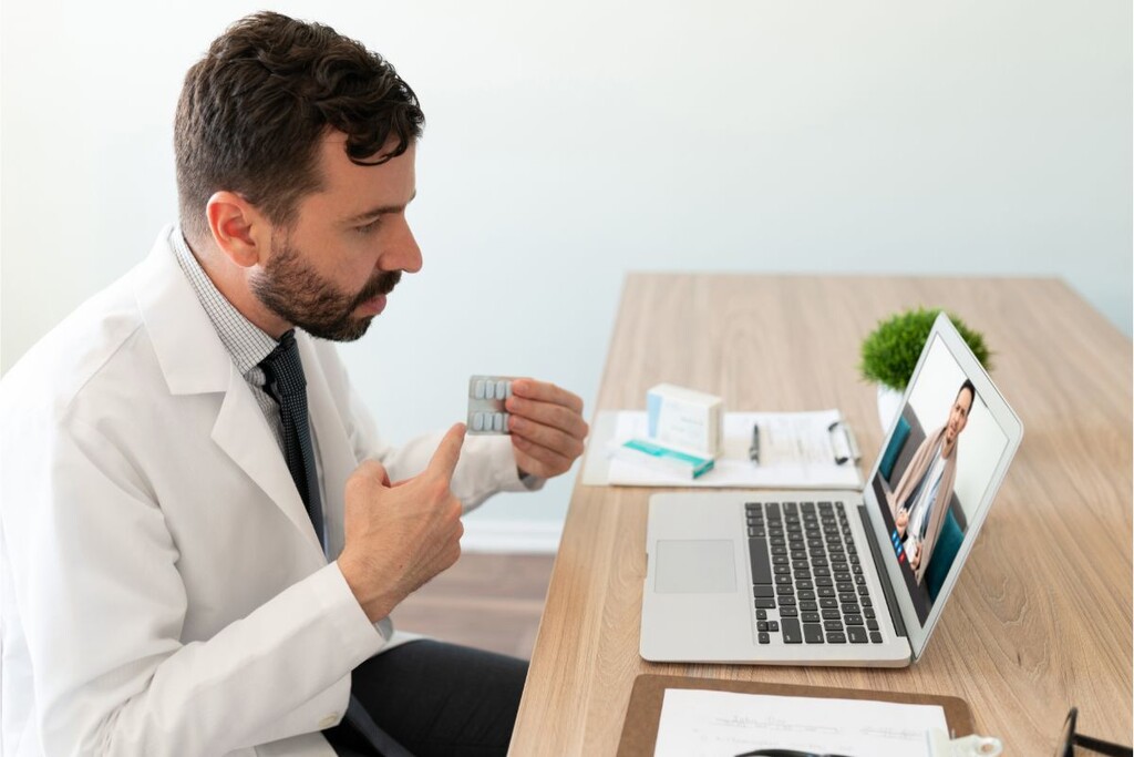 Médico de barba e jaleco apontando para cartela de remédios segurados por ele, em frente ao computador durante teleconsulta.