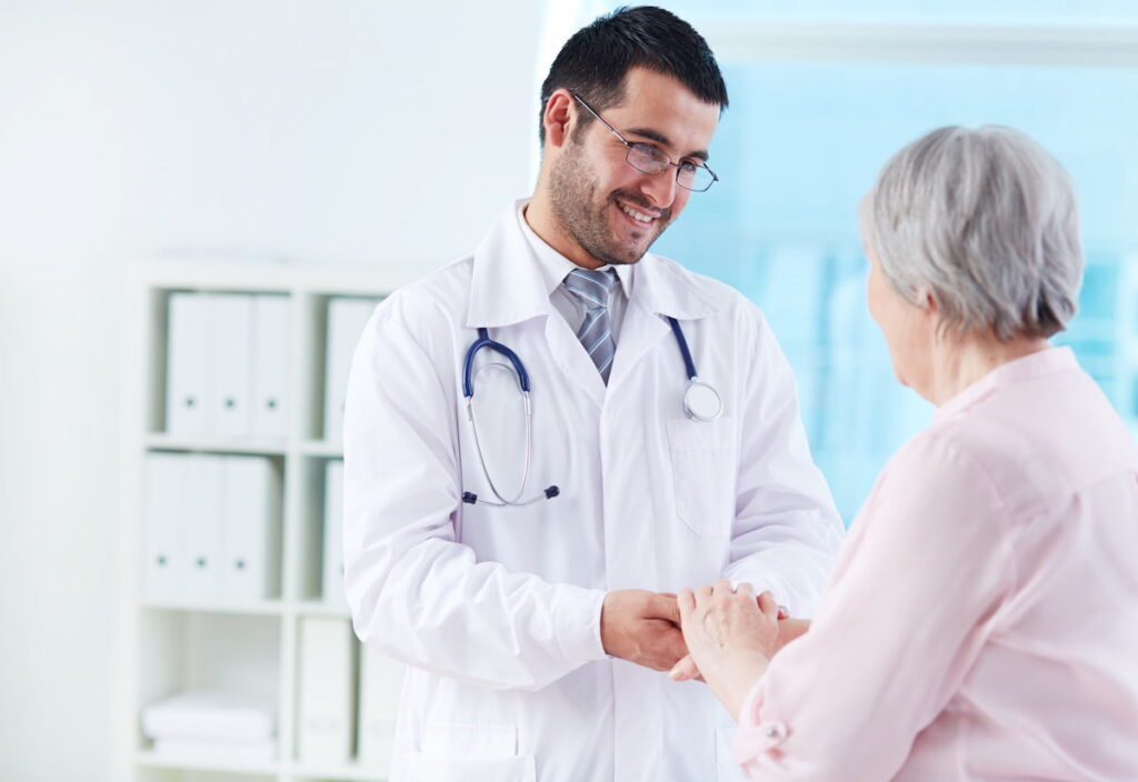 RQE médico: médico sorrindo e segurando a mão de uma idosa em um consultório.