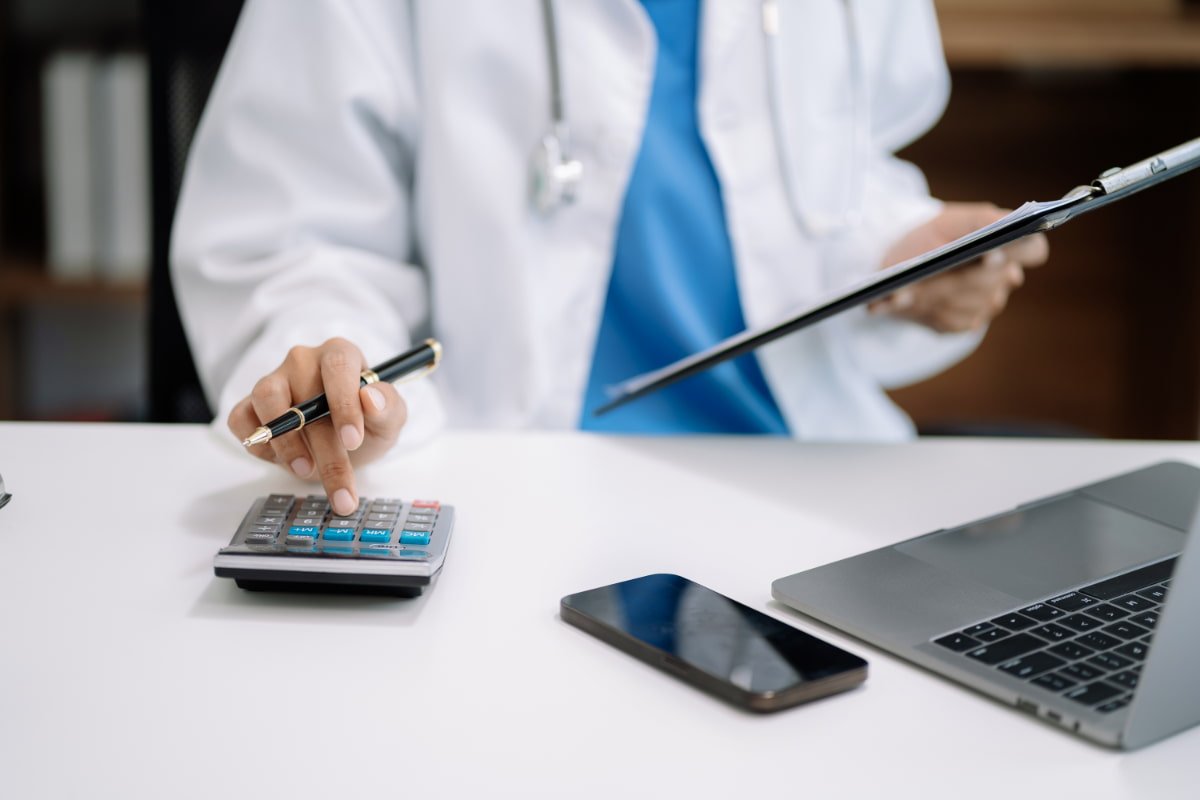Médico no seu consultório com uma calculadora, notebook e celular na mesa, aprendendo a como calcular o pró-labore médico.