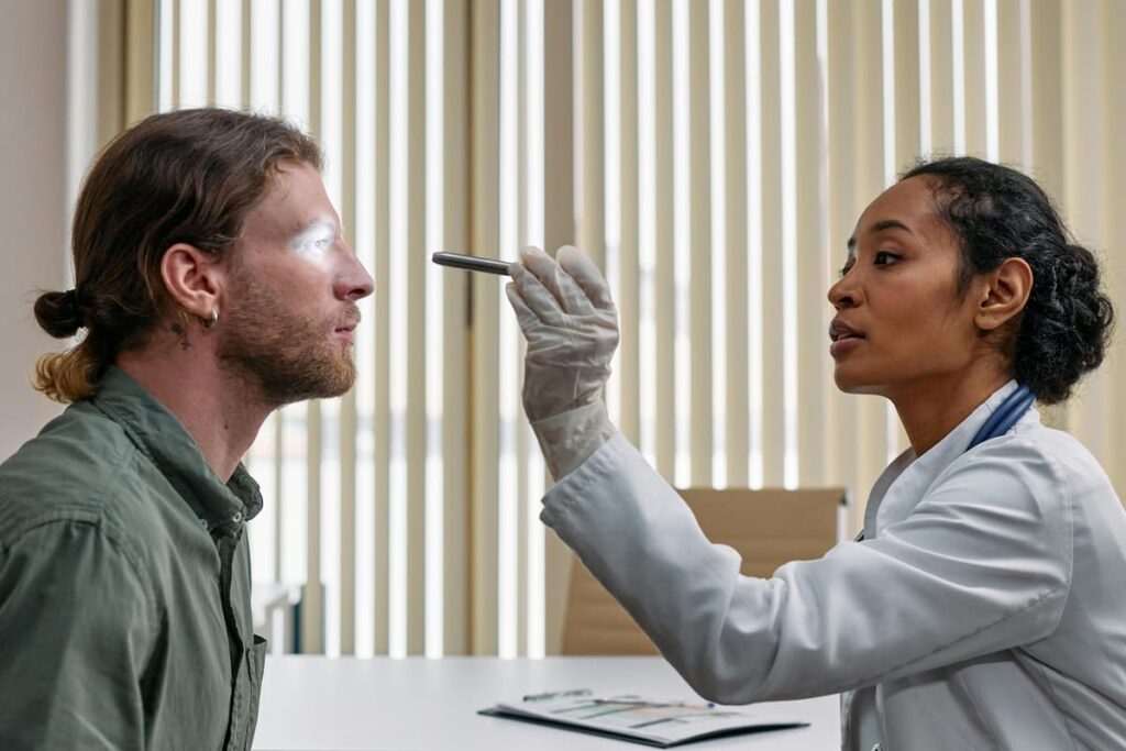 Em plantão médico, um profissional de saúde com jaleco branco examina os olhos de um paciente com uma lanterna clínica. Seguindo procedimentos do triple aim