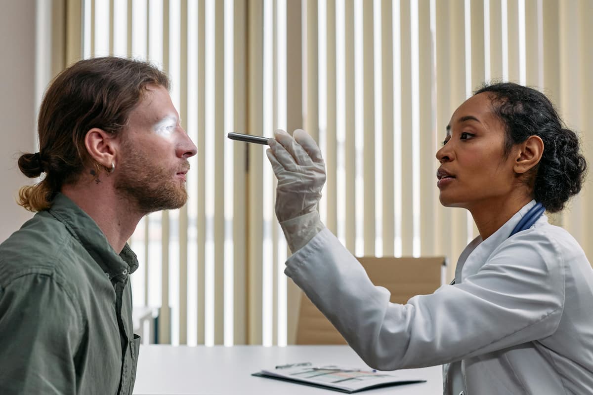 Em plantão médico, um profissional de saúde com jaleco branco examina os olhos de um paciente com uma lanterna clínica. Seguindo procedimentos do triple aim