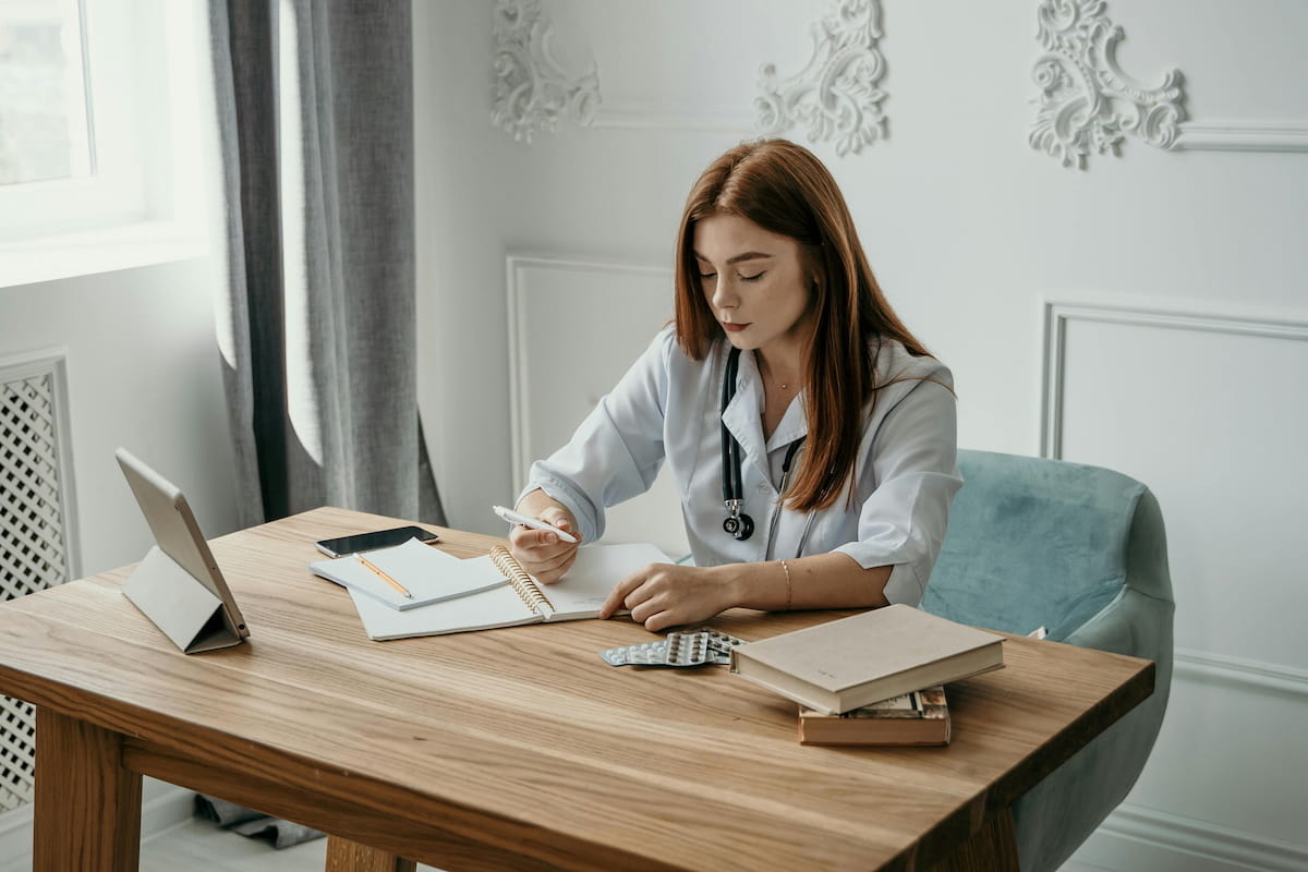 Médica preenchendo a DMED em um consultório, com laptop, papéis e estetoscópio sobre a mesa.