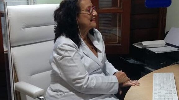 Médica de jaleco banco, sentada em uma cadeira também branca, em frente a um computador durante consulta.
