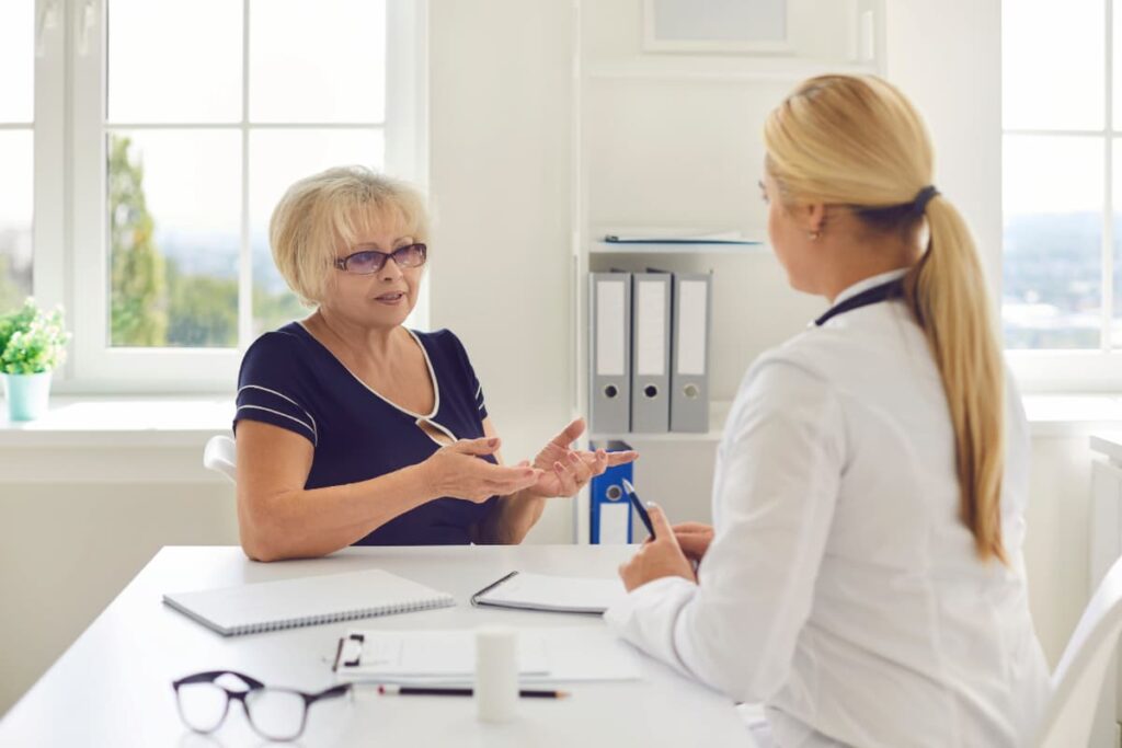 Paciente idosa em consulta de acompanhamento médico, conversando com uma profissional de saúde em um consultório iluminado.
