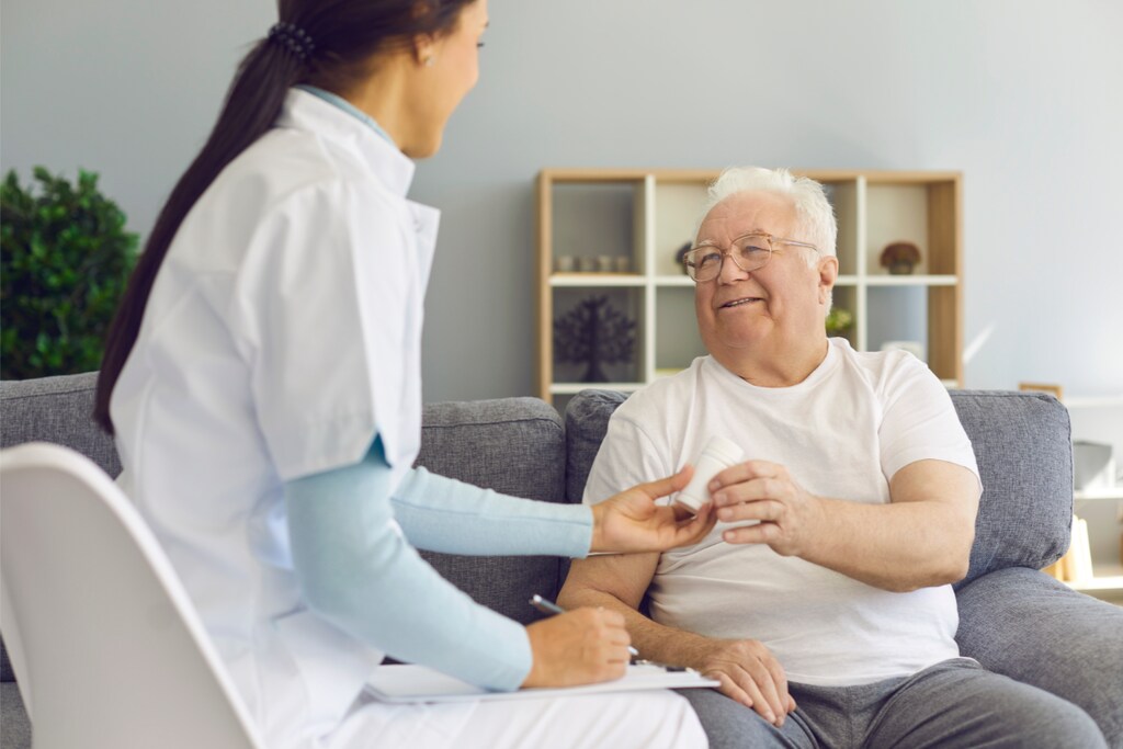 Um idoso sorridente recebe seus medicamentos de uma profissional de saúde em casa, representando o conceito de home care