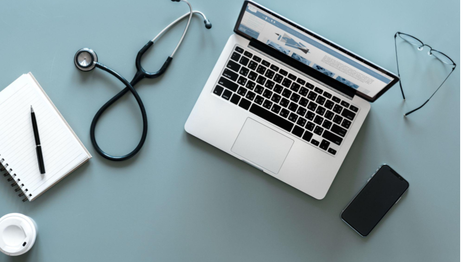 A imagem mostra uma cena organizada vista de cima, sobre uma superfície lisa em tom azul-acinzentado. No centro-direita, há um notebook aberto, com o teclado visível e a tela exibindo uma página com conteúdo textual e gráfico, sugerindo trabalho ou pesquisa online.