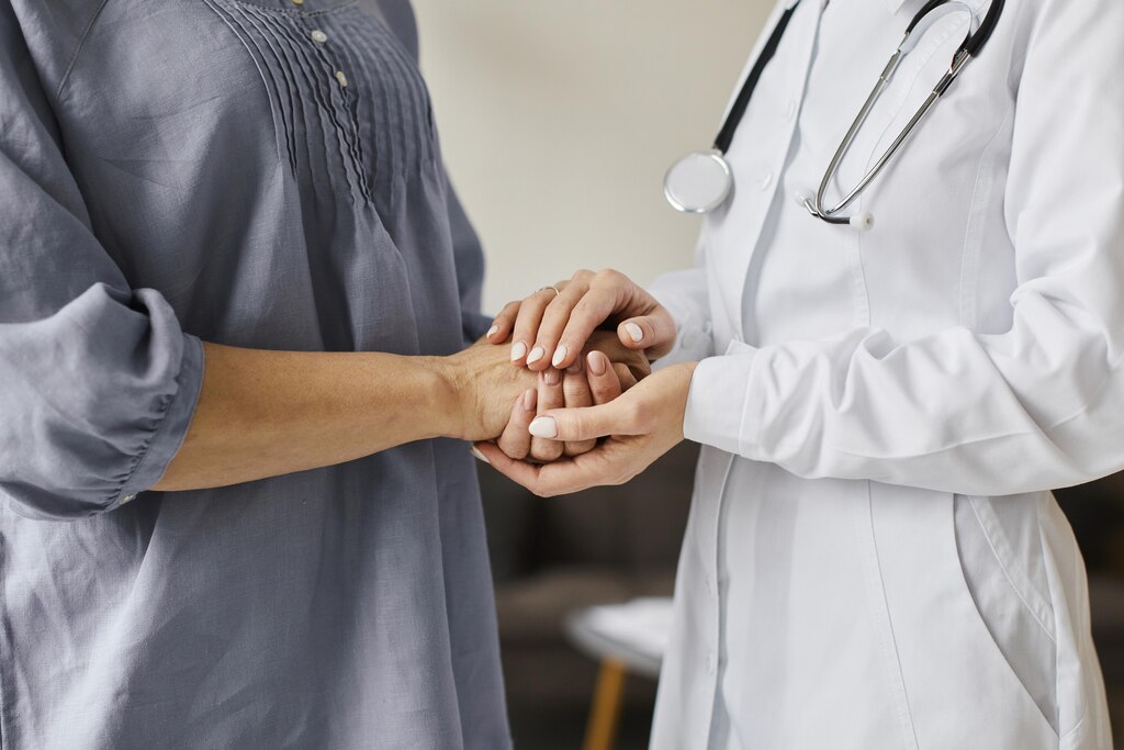 Médica, de jaleco branco, segurando as duas mãos do paciente de roupa cinza.