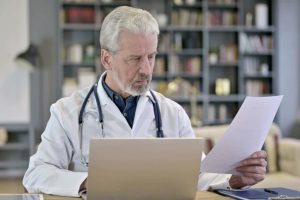 Médico olhando prontuário eletrônico do paciente (PEP) em papel com um computador na frente.