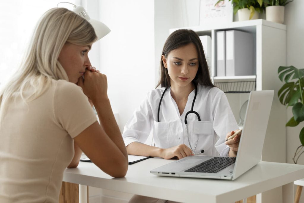 médica de jaleco branco faz uma anamnese completa junto com uma paciente, utilizando um notebook
