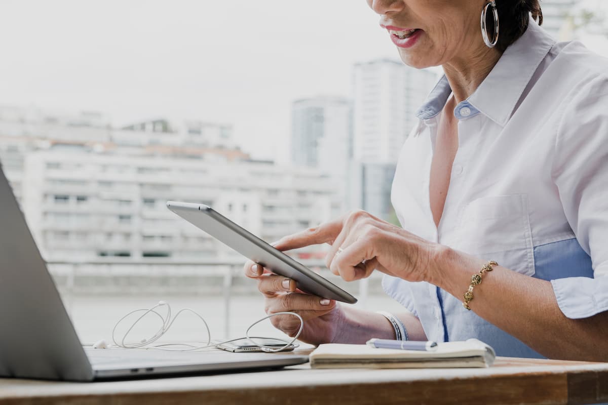 Mulher utilizando tablet ao lado de um laptop, representando tecnologia e agilidade na emissão de laudos a distância.