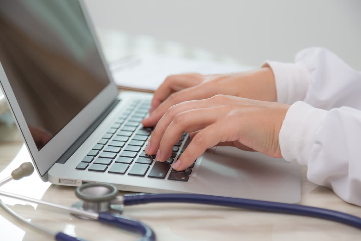 Médico digitando em laptop com estetoscópio ao lado, representando diferentes tipos de prontuário eletrônico na saúde.