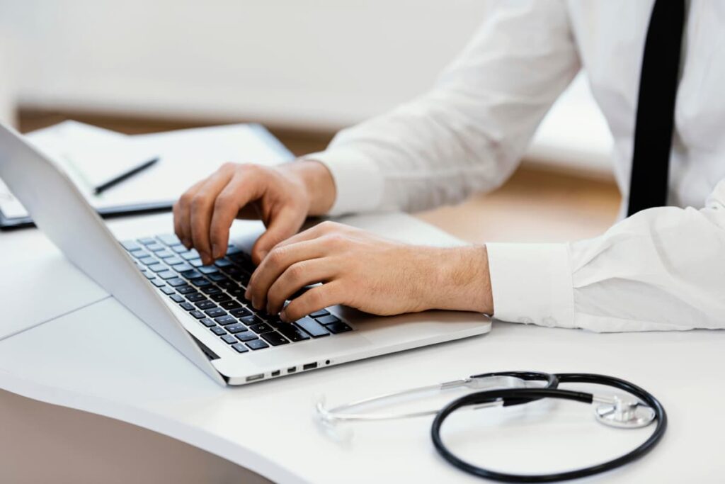 Médico digitando no laptop em um consultório, com estetoscópio sobre a mesa, representando o melhor software para controle financeiro.