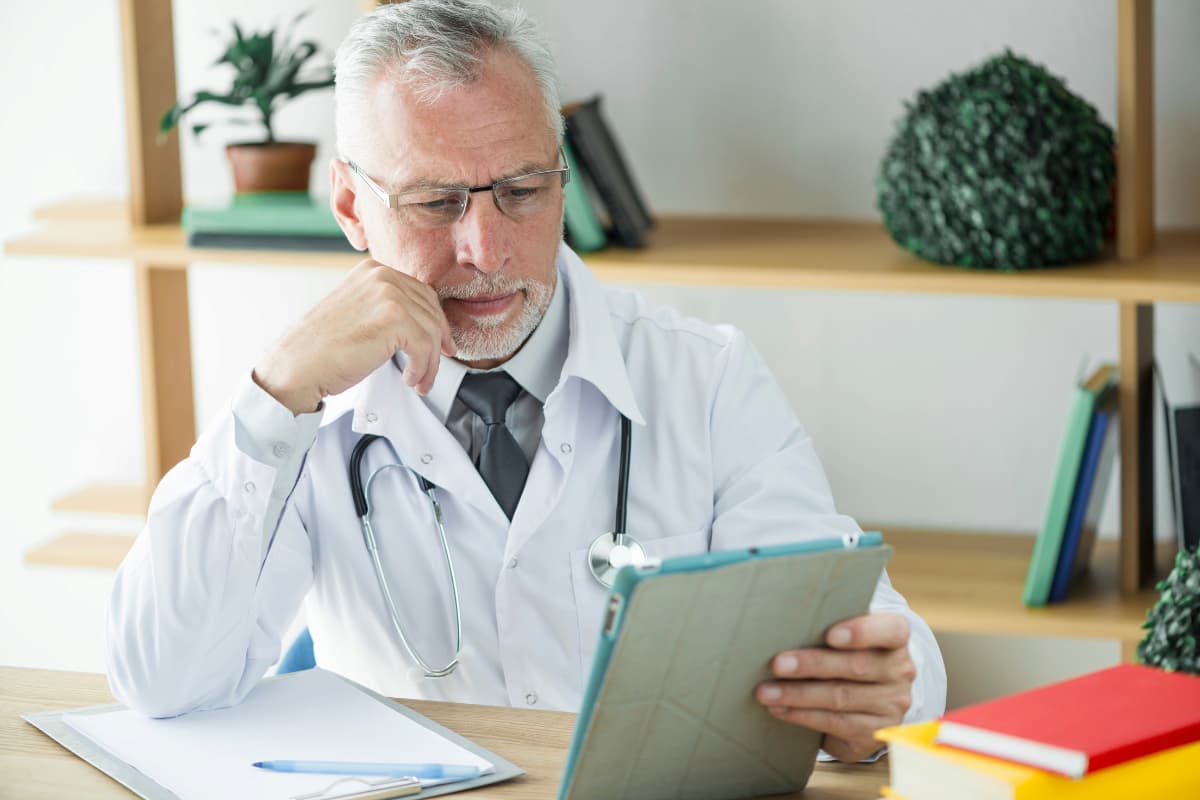 Homem de jaleco branco sentado à mesa, utilizando um tablet, representando saúde financeira para clínicas médicas.