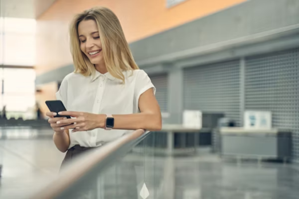 A imagem mostra uma mulher sorridente em um ambiente moderno e corporativo, usando um smartphone. Ela parece estar interagindo com um aplicativo, o que transmite praticidade, conectividade e satisfação com a experiência digital. Ela veste roupas sociais e usa um smartwatch, reforçando o perfil de alguém que valoriza eficiência e tecnologia no dia a dia.