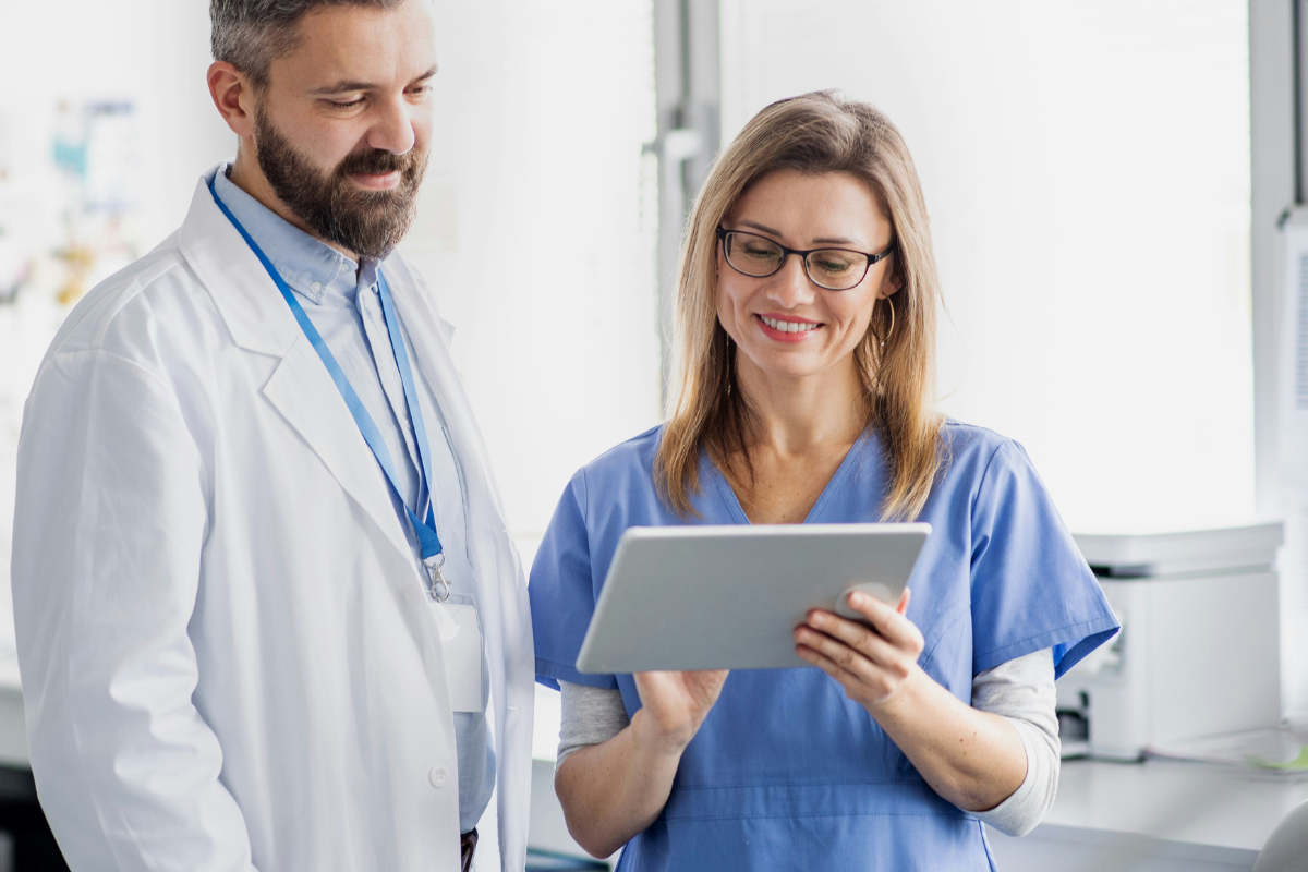A imagem mostra dois profissionais da saúde sorrindo enquanto olham para um tablet. Um homem com barba e jaleco branco (provavelmente um médico) está ao lado de uma mulher com óculos e vestida com roupa azul hospitalar (provavelmente uma enfermeira ou técnica de enfermagem). Ambos estão em um ambiente clínico ou hospitalar, com iluminação natural e equipamentos médicos ao fundo.