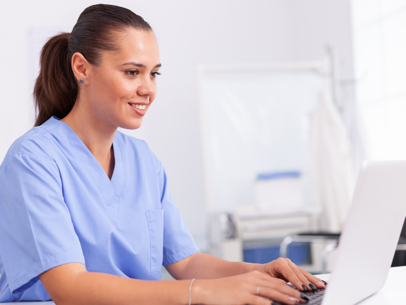 A imagem mostra uma profissional da saúde (provavelmente uma enfermeira ou técnica de enfermagem) sorrindo enquanto trabalha em um notebook em um ambiente clínico. Ela está vestida com roupa hospitalar azul clara e parece concentrada e satisfeita com o que vê na tela.