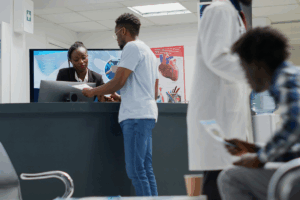 A imagem mostra o interior de uma clínica médica ou hospital. No centro da foto, um homem , vestindo uma camiseta cinza clara e jeans, está de pé em um balcão de recepção. Ele está interagindo com uma recepcionista , que está sentada atrás do balcão e parece estar examinando alguns papéis que o homem entregou.