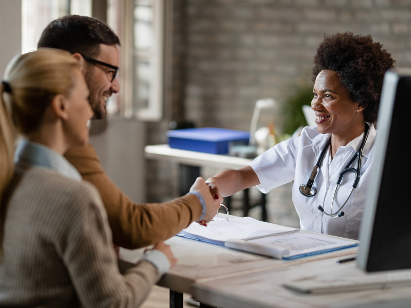 A imagem mostra uma médica, sorrindo e apertando a mão de um paciente do sexo masculino, em um consultório.