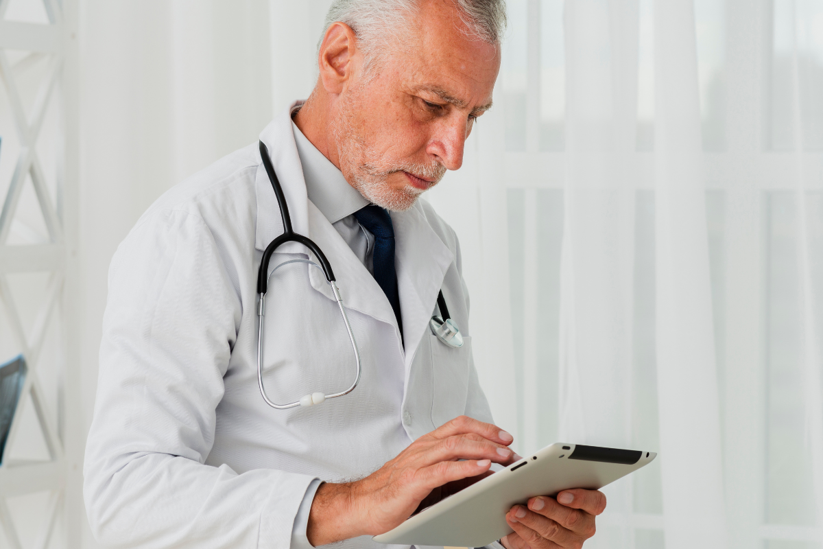 O médico está focado em um tablet (ou dispositivo similar) que segura com as duas mãos, parecendo estar lendo, analisando dados ou inserindo informações. Sua expressão é de concentração e seriedade.