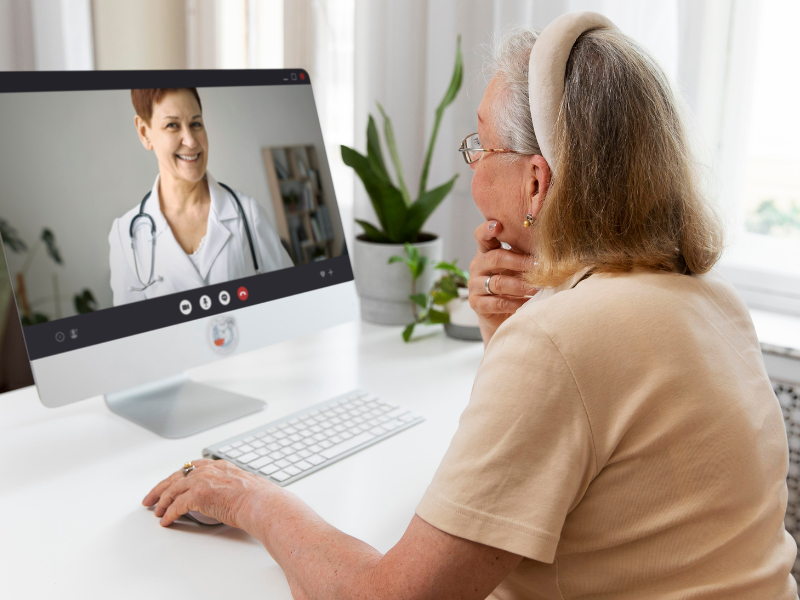 A imagem mostra uma mulher idosa participando de uma consulta médica online. Ela está sentada à frente de um computador de mesa, observando atentamente a tela, onde aparece uma profissional de saúde sorridente usando jaleco branco e um estetoscópio.