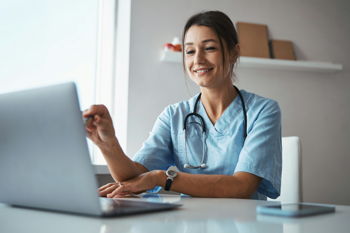 A imagem mostra uma profissional da área da saúde, vestindo um uniforme azul e com um estetoscópio pendurado no pescoço, sentada em uma mesa enquanto utiliza um laptop. Ela sorri e parece concentrada, interagindo com a tela do computador, possivelmente durante uma consulta online ou análise de dados médicos.