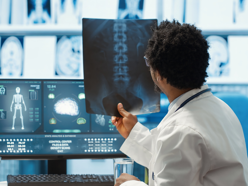 A imagem mostra um médico analisando uma radiografia em um ambiente clínico moderno, voltado para diagnóstico por imagem.