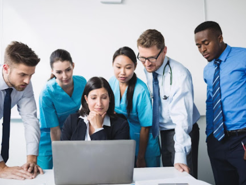A imagem mostra um grupo de seis profissionais, incluindo médicos e pessoal administrativo, reunidos em torno de um laptop para analisar ou discutir informações importantes.