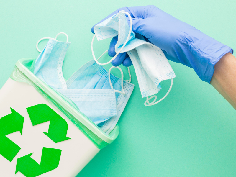 A imagem mostra uma mão com luva descartável azul segurando várias máscaras cirúrgicas usadas e depositando-as em uma lixeira branca com o símbolo verde de reciclagem estampado na frente.