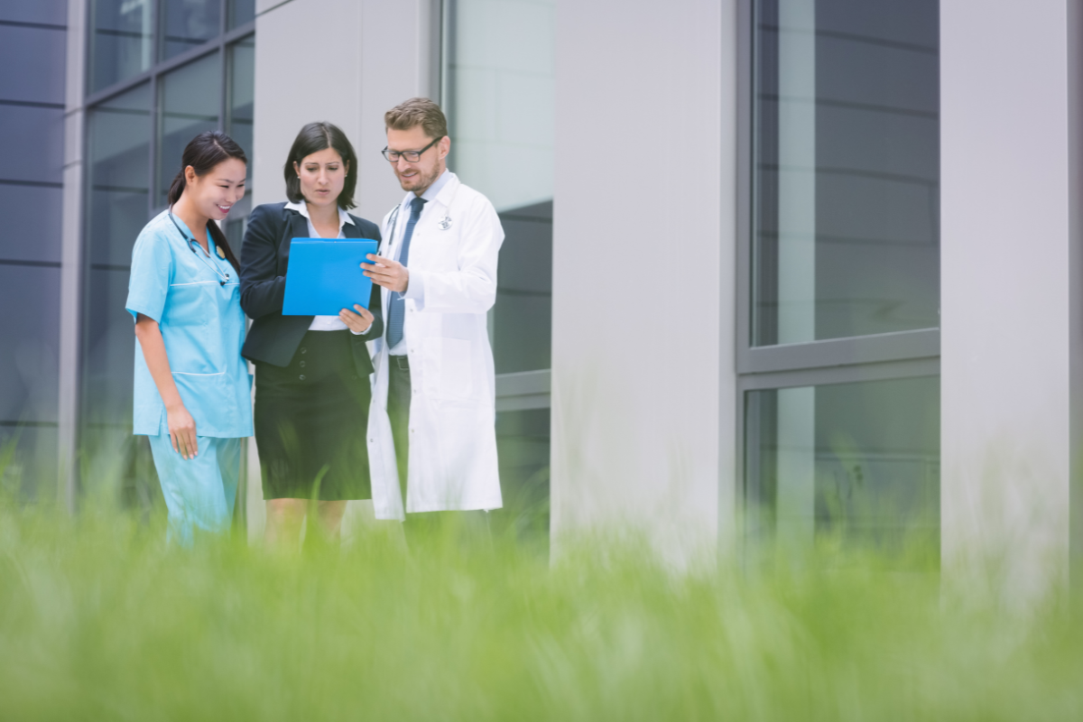 A imagem mostra três profissionais conversando em um ambiente externo de um prédio corporativo ou hospitalar, transmitindo colaboração e alinhamento entre equipes da área da saúde.