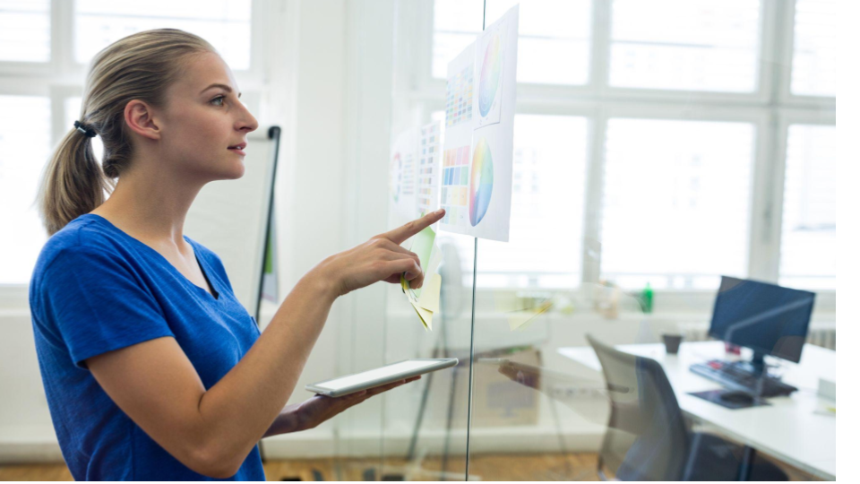 A imagem mostra uma mulher em um ambiente corporativo ou de escritório moderno, de perfil, interagindo com um painel de vidro transparente onde estão fixados gráficos e diagramas coloridos, como gráficos de pizza e tabelas.