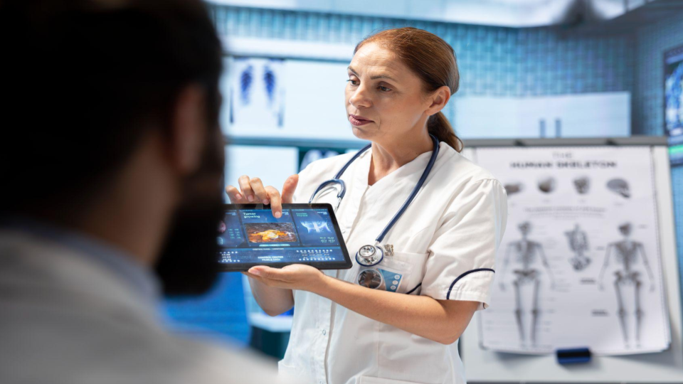 A imagem mostra uma profissional de saúde, provavelmente uma médica ou enfermeira, usando uniforme branco e estetoscópio no pescoço, explicando informações a um paciente em um ambiente clínico.