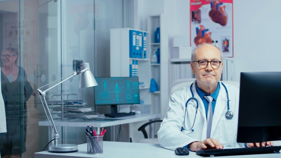 A imagem mostra um médico sentado em seu consultório, trabalhando em frente a um computador. Ele usa jaleco branco, estetoscópio no pescoço e óculos, e olha para a câmera com uma expressão calma e profissional, transmitindo confiança e experiência.