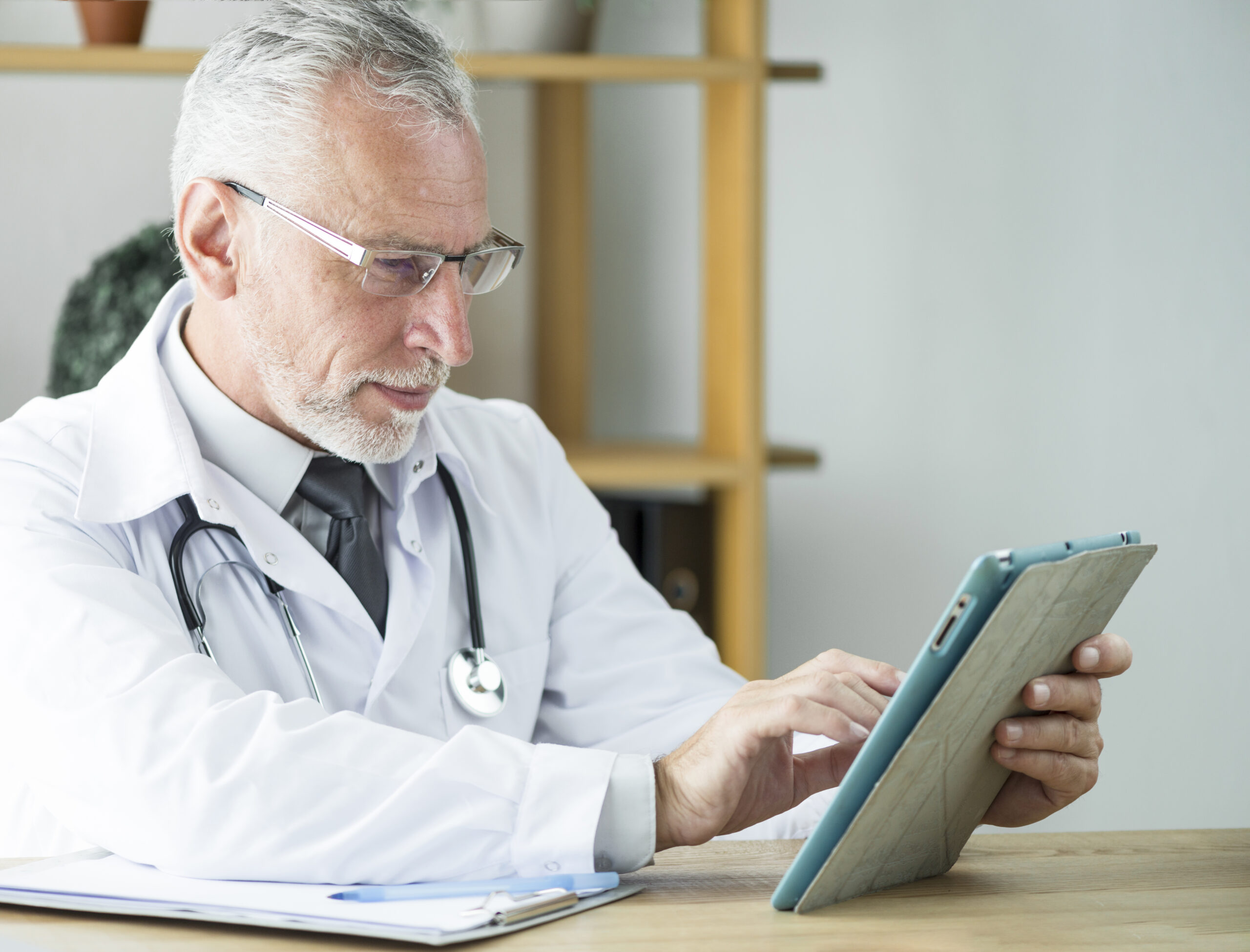 Esta imagem apresenta um homem, um médico sênior, com cabelo e barba grisalhos, com cerca de 60 anos. Ele está sentado em uma mesa de madeira, concentrado em seu trabalho.