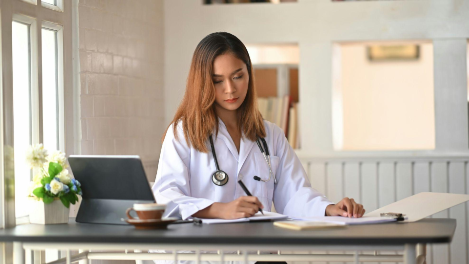 A imagem mostra uma profissional da área da saúde, possivelmente uma médica, sentada em uma mesa enquanto escreve em um caderno ou ficha.