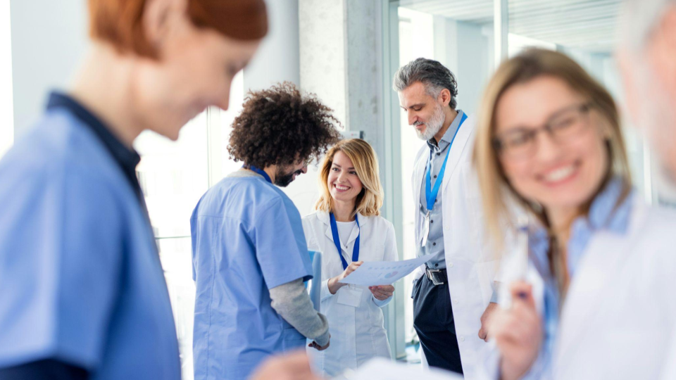 A imagem retrata um grupo de profissionais de saúde em um ambiente moderno, bem iluminado e de aspecto clínico (como um hospital ou centro de pesquisa). A composição foca na colaboração e no dinamismo da equipe.