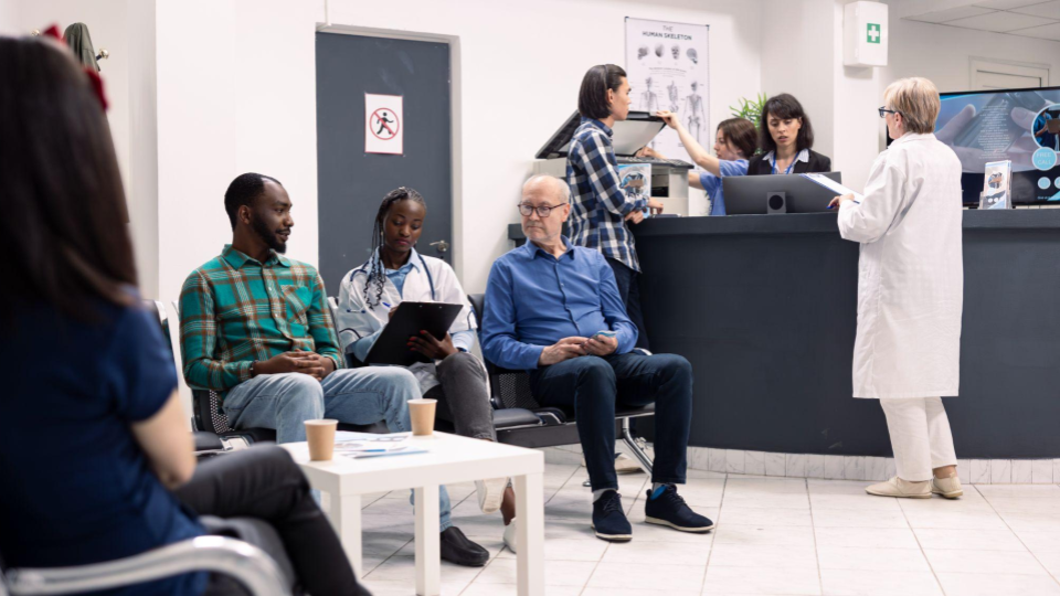 A imagem retrata a rotina na sala de espera e na recepção de uma clínica médica ou hospital. A cena mostra diferentes interações acontecendo simultaneamente em um ambiente bem iluminado.