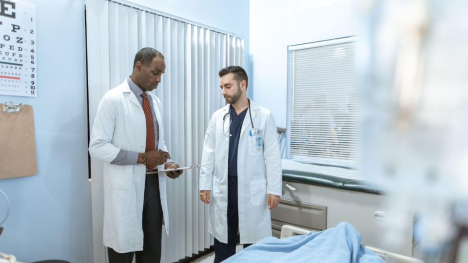 A imagem retrata dois médicos conversando no que parece ser um quarto de hospital ou uma sala de exames clínicos. O ambiente transmite uma atmosfera profissional, colaborativa e de cuidado médico.