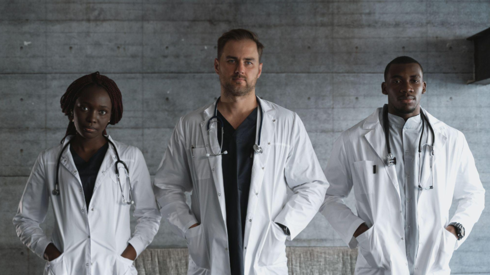 A imagem mostra um retrato de um grupo diversificado composto por três profissionais da saúde (médicos ou enfermeiros), posando de forma confiante e séria.