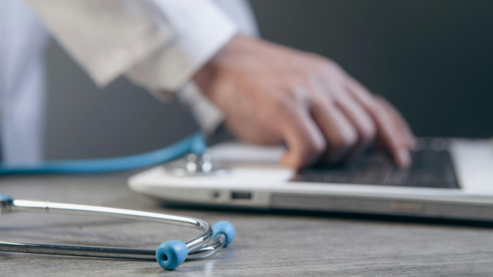 Uma imagem com desfoque artístico que destaca as ferramentas de trabalho digitais e analógicas de um médico.
