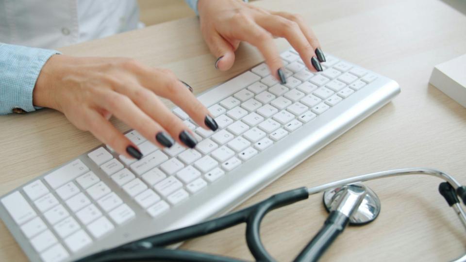 Uma visão superior diagonal focada nas mãos de uma profissional de saúde inserindo dados no computador.