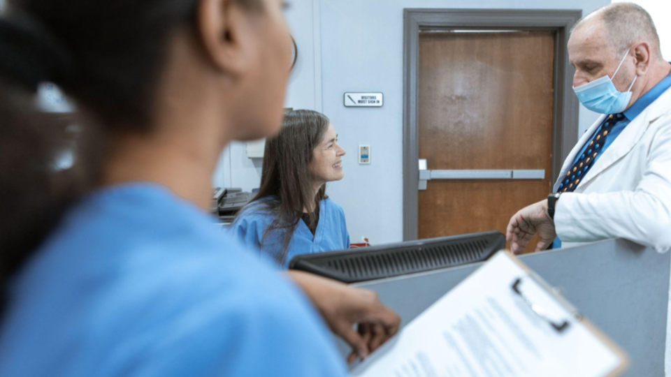 A imagem mostra um ambiente médico ou hospitalar, focado na interação entre profissionais de saúde em um balcão ou recepção.