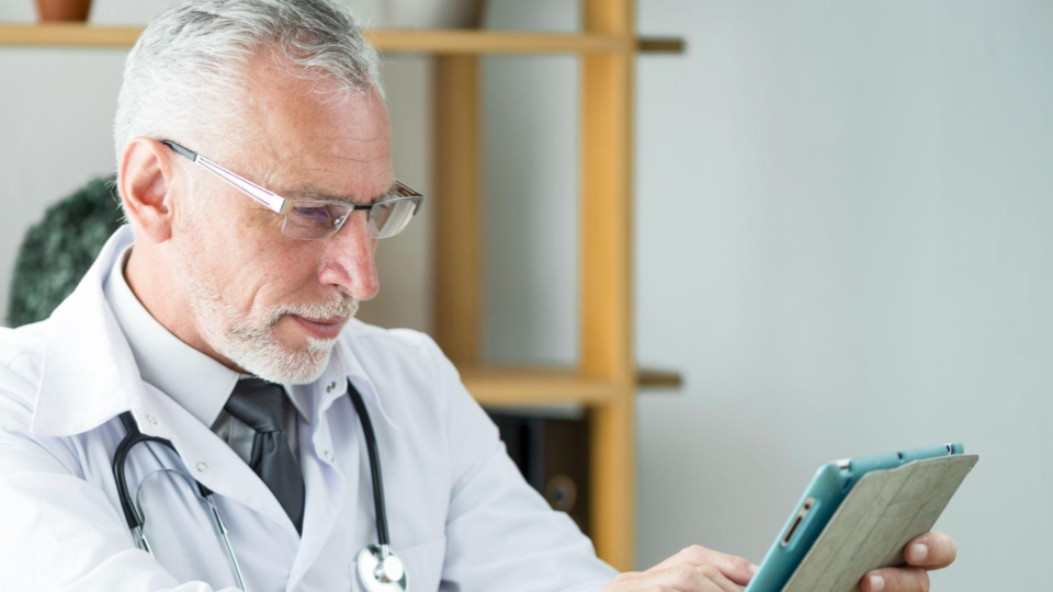 A imagem mostra um médico sênior concentrado enquanto utiliza um tablet.