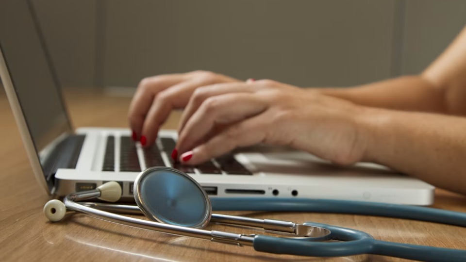 Plano detalhado das mãos de uma profissional de saúde digitando em um notebook de tom prateado, com um estetoscópio posicionado em primeiro plano sobre a mesa de madeira.