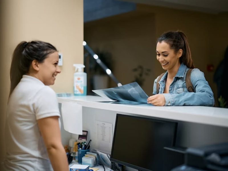 Esta imagem mostra uma cena de atendimento em recepção, possivelmente em uma clínica ou consultório. À esquerda, uma profissional (possivelmente recepcionista) sorri enquanto interage com uma jovem mulher do outro lado do balcão, que também sorri enquanto segura exames de imagem, como raio-x.